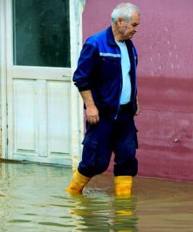 An older man wearing yellow boots walking through a flooded area near a building. - Olive Oil Times