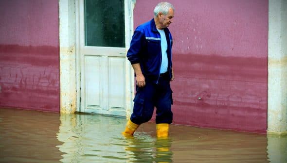 An older man wearing yellow boots walking through a flooded area near a building. - Olive Oil Times