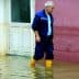 An older man wearing yellow boots walking through a flooded area near a building. - Olive Oil Times
