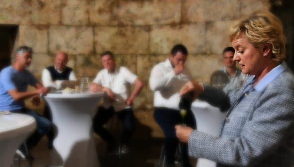 A woman in a gray blazer pouring olive oil while a group of men observes at a tasting event. - Olive Oil Times