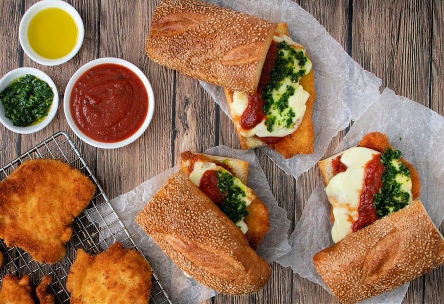 Two Italian sandwiches filled with fried chicken, cheese, and sauces on a wooden surface. - Olive Oil Times