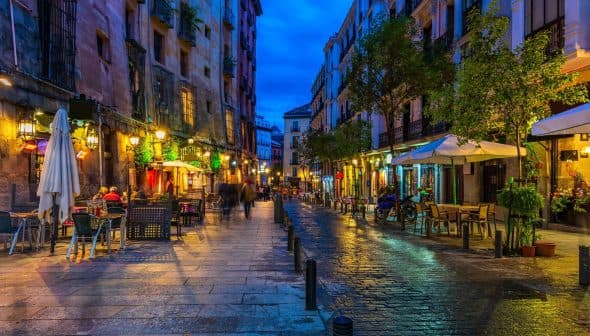 Narrow street in a European city illuminated by lights with outdoor dining areas and wet cobblestones. - Olive Oil Times