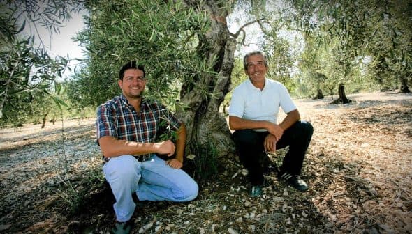 Two men sitting beside an olive tree in an olive grove, both smiling at the camera. - Olive Oil Times