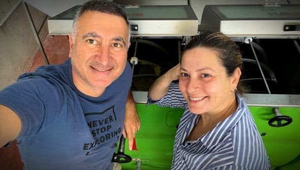 Two individuals smiling while standing next to machinery in a workshop environment. - Olive Oil Times