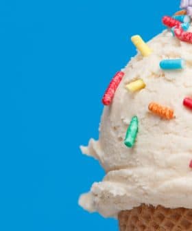 Close-up of an ice cream cone topped with vanilla ice cream and colorful sprinkles against a blue background. - Olive Oil Times
