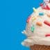 Close-up of an ice cream cone topped with vanilla ice cream and colorful sprinkles against a blue background. - Olive Oil Times
