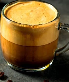 A glass mug filled with coffee featuring a distinct layer of cream on top, surrounded by coffee beans. - Olive Oil Times