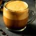 A glass mug filled with coffee featuring a distinct layer of cream on top, surrounded by coffee beans. - Olive Oil Times