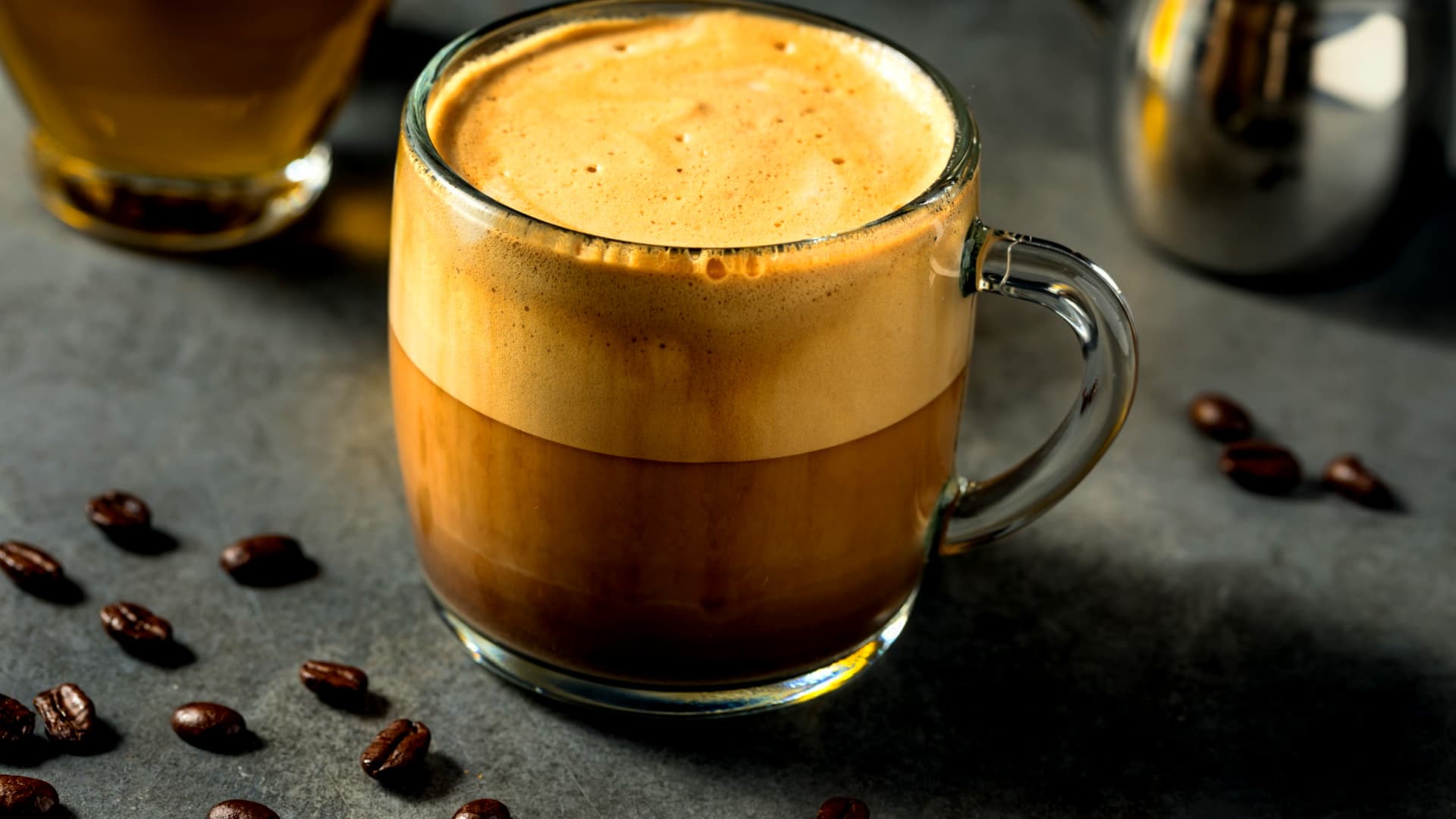 A glass mug filled with coffee featuring a distinct layer of cream on top, surrounded by coffee beans. - Olive Oil Times