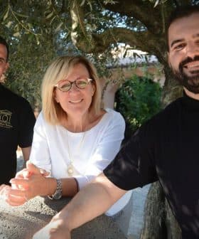 Three people standing together outdoors, smiling and posing for a photo near an olive tree. - Olive Oil Times