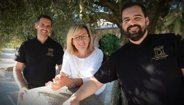 Three people standing together outdoors, smiling and posing for a photo near an olive tree. - Olive Oil Times