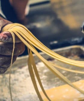 A hand holding freshly rolled pasta strands from a pasta machine in a kitchen setting. - Olive Oil Times