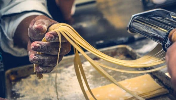 A hand holding freshly rolled pasta strands from a pasta machine in a kitchen setting. - Olive Oil Times
