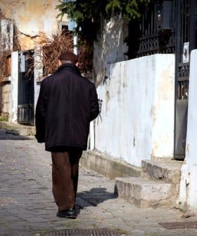 An elderly man walking away on a cobblestone street lined with buildings and greenery. - Olive Oil Times