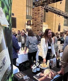 A booth displaying olive oil products at an exhibition with people interacting in the background. - Olive Oil Times