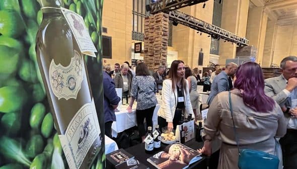 A booth displaying olive oil products at an exhibition with people interacting in the background. - Olive Oil Times