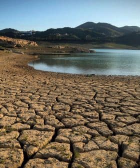 Dry, cracked earth surrounding a small body of water with mountains in the background. - Olive Oil Times
