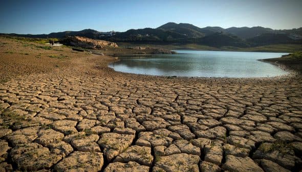 Dry, cracked earth surrounding a small body of water with mountains in the background. - Olive Oil Times