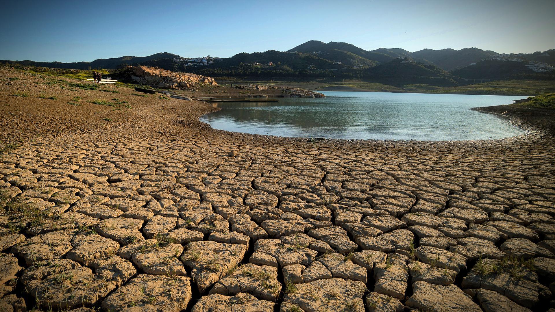 Dry, cracked earth surrounding a small body of water with mountains in the background. - Olive Oil Times