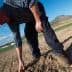 A farmer in a plaid shirt and hat kneeling down to inspect the soil in a field. - Olive Oil Times