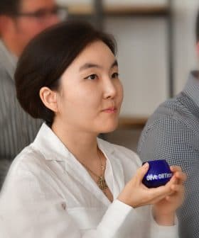 A woman with short hair holding a blue container while seated among a group of people. - Olive Oil Times