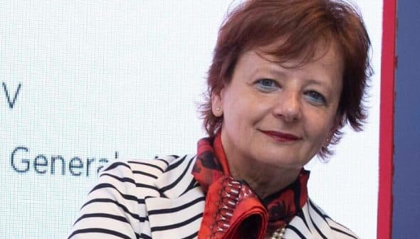 Woman with short red hair wearing a striped blazer and a red scarf, posing for the camera. - Olive Oil Times