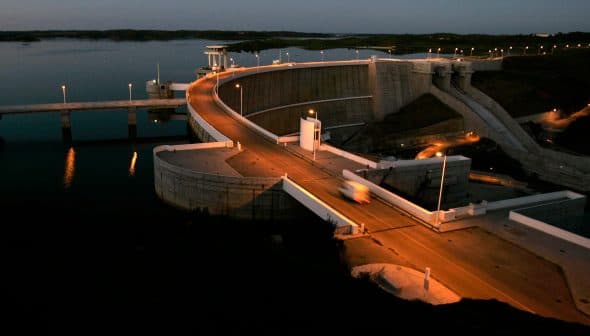 Concrete dam structure with a roadway and lights at dusk, reflecting in the water below. - Olive Oil Times