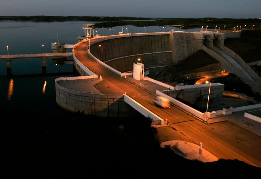 Concrete dam structure with a roadway and lights at dusk, reflecting in the water below. - Olive Oil Times