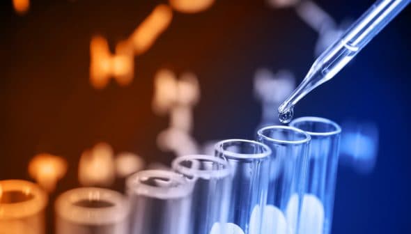 A pipette dropping liquid into a row of test tubes with a blurred clock in the background. - Olive Oil Times