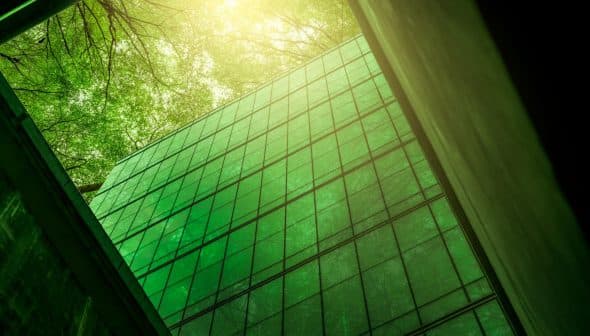 A modern glass building reflecting green light from surrounding trees in an urban setting. - Olive Oil Times