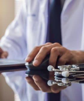 A medical professional in a white coat using a laptop with a stethoscope on the table. - Olive Oil Times