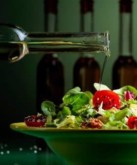 A hand pouring olive oil from a glass bottle onto a salad in a green bowl with cherry tomatoes and greens. - Olive Oil Times