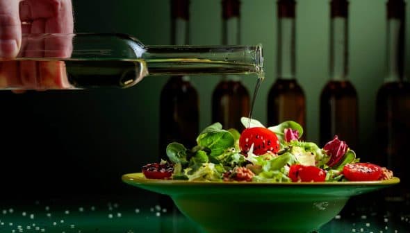 A hand pouring olive oil from a glass bottle onto a salad in a green bowl with cherry tomatoes and greens. - Olive Oil Times