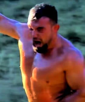 A shirtless man with a muscular build raising his arm in celebration outdoors. - Olive Oil Times