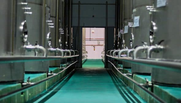 Interior view of a processing facility with stainless steel oil storage tanks and pipes. - Olive Oil Times