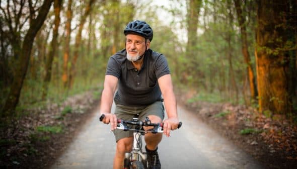 Older man wearing a helmet riding a bicycle on a forest trail during daylight. - Olive Oil Times