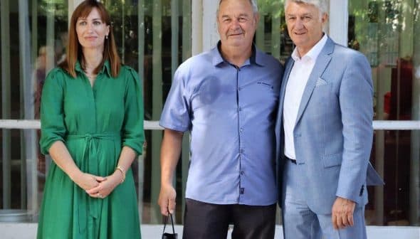 Three individuals standing together for a photo, with one woman in a green dress and two men in casual and formal attire. - Olive Oil Times