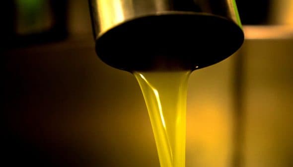 Golden olive oil flowing from a metal tap into a container in a close-up view. - Olive Oil Times