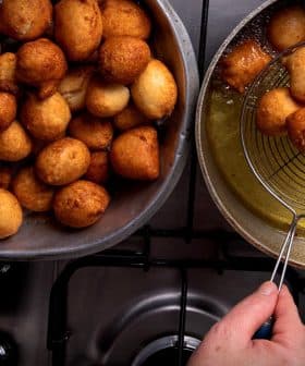 A bowl filled with golden-brown fried dough balls next to a frying pan with more dough balls being drained. - Olive Oil Times