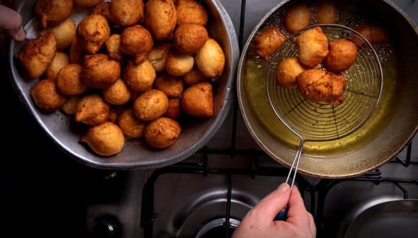 A bowl filled with golden-brown fried dough balls next to a frying pan with more dough balls being drained. - Olive Oil Times