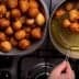 A bowl filled with golden-brown fried dough balls next to a frying pan with more dough balls being drained. - Olive Oil Times