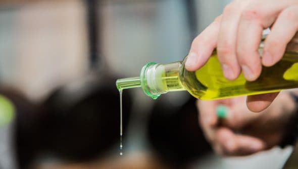 A person pouring olive oil from a glass bottle with a green cap, with oil dripping from the spout. - Olive Oil Times