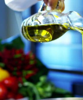 A person pouring olive oil from a glass bottle into a metal bowl in a kitchen setting. - Olive Oil Times