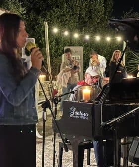 A woman singing into a microphone while seated at a piano during a performance at night. - Olive Oil Times