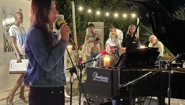 A woman singing into a microphone while seated at a piano during a performance at night. - Olive Oil Times