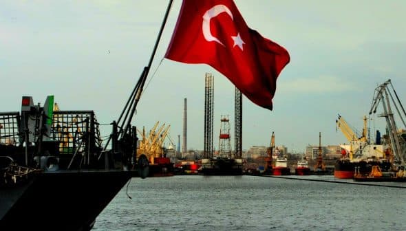 Turkish flag waving on a ship with cranes and industrial structures in the background. - Olive Oil Times