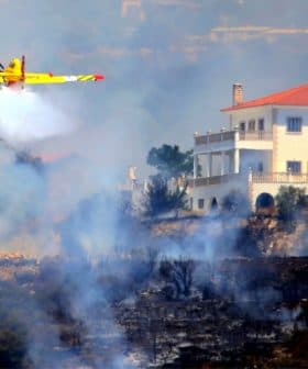 Yellow firefighting airplane releasing water over a wildfire near a white house. - Olive Oil Times