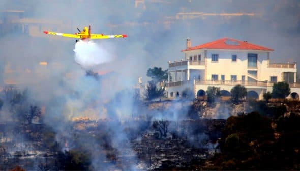 Yellow firefighting airplane releasing water over a wildfire near a white house. - Olive Oil Times