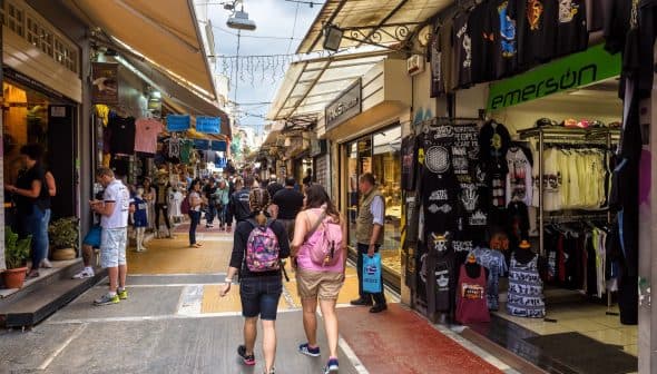 Two people walking through a market street lined with shops and stalls selling various items. - Olive Oil Times