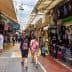 Two people walking through a market street lined with shops and stalls selling various items. - Olive Oil Times